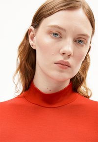 Rotes Oberteil mit hohem Hals aus glattem Stoff. Nahaufnahme einer Person mit welligem, hellbraunem Haar, die kleine Ohrringe trägt und einen neutralen Gesichtsausdruck hat.