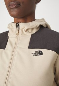 Beige a tmavě šedá zipová bunda s kapucí, se znakem na hrudi a texturovanou tkaninou. Výrazný design s barevnými bloky.