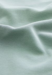 Lichtgroene katoenen stof met een gladde textuur, voorzien van kleine, subtiele geweven patronen. De stof hangt zacht en heeft kreukjes.