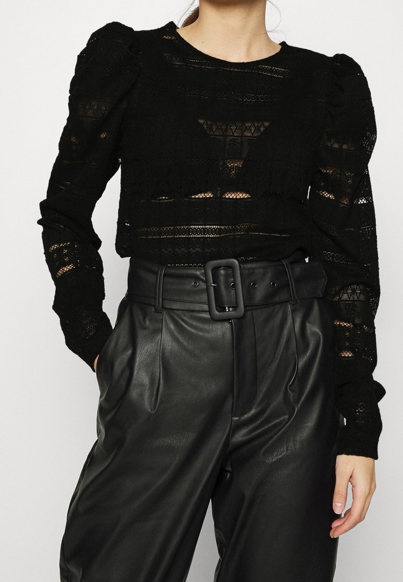 Femme portant un haut noir en dentelle à manches longues avec des épaules bouffantes et un pantalon en cuir noir taille haute avec une large ceinture et une boucle rectangulaire.