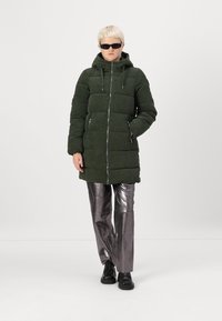 Abrigo tipo puffer de pana verde con cierre en la parte frontal, capucha ajustable y dos bolsillos laterales; combinado con pantalones metálicos brillantes y zapatos negros.