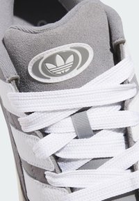 Grey suede sneaker with white leather stripes, featuring a grey logo patch and white laces. Close-up of the upper section and lace area.