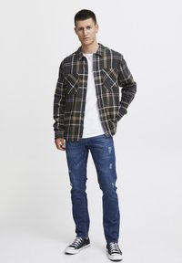 Geruite flanellen shirt met donkere groene, zwarte en beige kleuren, lange mouwen, twee borstzakken, gecombineerd met versleten blauwe jeans en zwarte sneakers.
