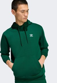 Zelená mikina s kapucňou vyrobená z mäkkého materiálu, s prednou vreckom, šnúrkou na stiahnutie kapucne a bielym logom Adidas na hrudi. Jednoduchý dizajn, voľný strih.