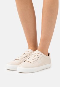 Baskets en cuir beige avec semelles en caoutchouc blanches, présentant un design à lacets et un marquage subtil sur le côté. Texture lisse, style basse.