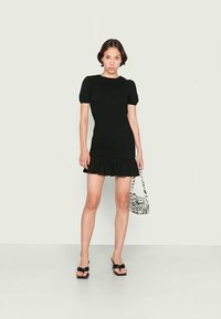 Vestido mini preto ajustado com mangas curtas em balão, barra com babados, combinado com uma mala de zebra e chinelos pretos em um fundo claro.