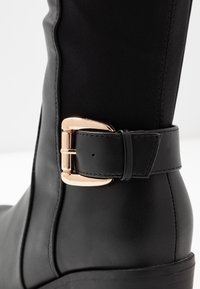 Botín negro hasta la rodilla con una parte superior de cuero suave, detalle de hebilla en tono dorado y una correa texturizada. Las costuras cosidas añaden interés estructural.