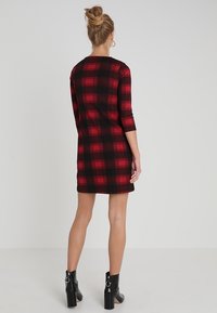 Šaty do délky kolen v černé a červené kostičce se třičtvrtinovými rukávy. Nosené s černými kotníkovými botami s stříbrným akcentem. Hladká textilie.