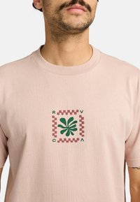Lichtroze t-shirt van katoen met een centrale afbeelding van een groen bladontwerp en een bruine geruite rand. Ronde hals en korte mouwen.