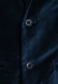 Fluwelen blazer in donker marineblauw, met twee zwarte, gelabelde knopen. De stof heeft een gladde textuur met een subtiele glans.