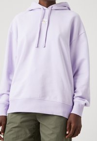 Felpa oversized di un leggero viola con tasca a canguro frontale, cappuccio con coulisse e polsini a coste, indossata sopra pantaloni verde oliva.