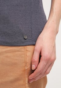 Chemise grise à manches courtes avec une texture douce, présentant un petit accent logo métallique près de l'ourlet. Main touchant la chemise, avec un pantalon beige visible.