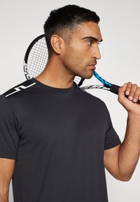 Svart fukttransporterande träningströja med rund halsringning, med vita detaljer på axeln, hålls mot en tennisracket.