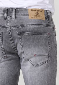 Šedé džíny z denimu s vybledlým vzhledem, designem s pěti kapsami a červenými švy. Nápis na pásku přečtete "Rugged Denim."