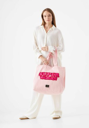 Roze canvas draagtas met "SUPER WOMAN" in vette, fuzzy letters. Voorzien van stevige hanen en een klein merklabel aan de onderkant. Ideaal voor casual gebruik.
