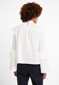 Vit blus med volangkrage och trumpetärmar, prydd med ett strukturerat prickmönster. Baksidan har en nyckelhålsknäppning i nacken.