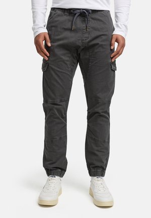 Hombre con pantalones jogger cargo gris oscuro con cintura ajustable y zapatillas blancas, de pie contra un fondo liso.