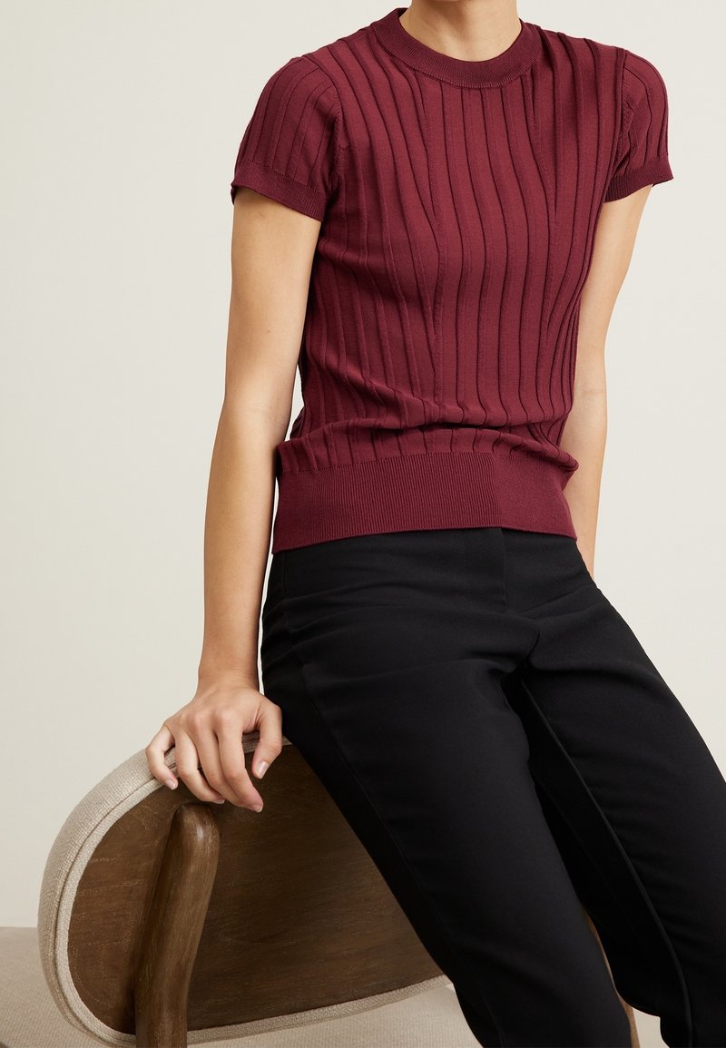 Personne portant un haut côtelé bordeaux à manches courtes et un pantalon noir, assise de côté sur une chaise beige en bois, la main reposant sur le dossier de la chaise.