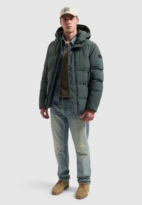 Donkergroene gewatteerde jas met een capuchon, gelaagd over een spijkerhemd en een olijfgroen vest. Lichtblauwe versleten jeans en tan schoenen maken de outfit compleet.