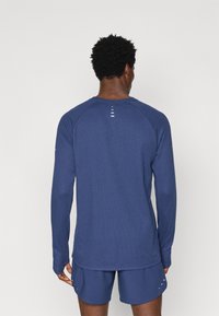 Μπλούζα αθλητική με μακριά μανίκια σε ναυτική απόχρωση, με υφή, ράγλαν μανίκια και ανακλαστικές λεπτομέρειες στο πίσω μέρος για αυξημένη ορατότητα.