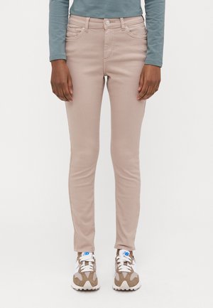 Person iført beige slim-fit jeans, teal langærmet top og beige og hvide sneakers med blå logoer på hvid baggrund.