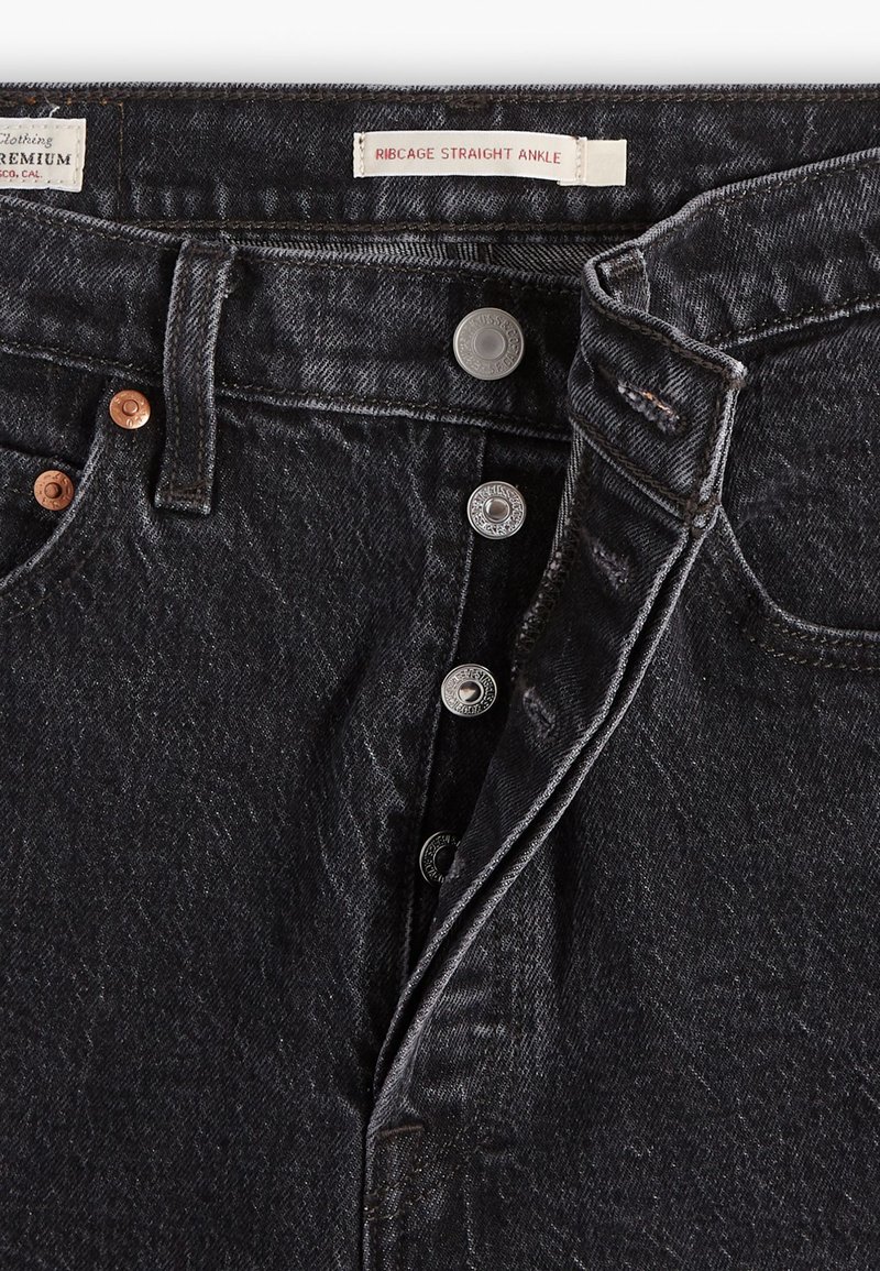 Jean en denim noir avec une coupe droite à la cheville, une fermeture à cinq boutons, et des accents de quincaillerie en cuivre et argent. Des détails de texture et de couture subtils sont visibles.