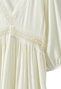 Abito color crema con rifiniture in pizzo floreale lungo il collo a V e in vita, caratterizzato da un tessuto texturizzato e dettagli di gonna arricciata.