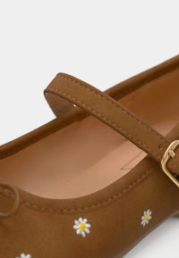 Καφέ μπαλαρίνα με στρογγυλεμένη μύτη, μονή τιράντα και χρυσή αγκράφα. Διακοσμημένη με λευκά και κίτρινα κεντητά μαργαριτάρια στο πλάι. Λείο υλικό.