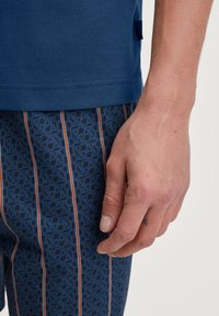 Navy blauwe kortemouwen shirt gecombineerd met gestreepte broek met patronen van herhalende logo's in blauw en oranje. Close-up van een hand die op de stof rust.