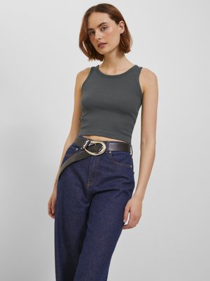 Kobieta z krótkimi brązowymi włosami, ubrana w ciemnoszary bezrękawnik typu crop top, niebieskie jeansy z wysokim stanem oraz czarny pasek z dużą srebrną sprzączką.
