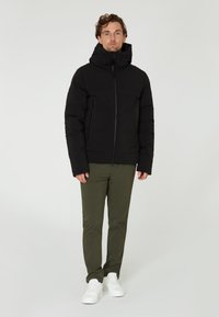 Chaqueta acolchada negra con cuello alto y capucha desmontable, con dos bolsillos frontales con cremallera. Combinada con pantalones verde olivo y zapatillas blancas.