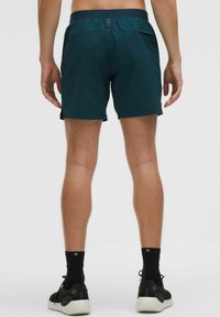 Tyrkizkék sportnadrág sima textúrával, rugalmas derékpánttal és oldalsó hasítékokkal a mozgékonyság érdekében. Fekete zoknikkal és tornacipővel viselve.