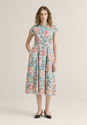 Kvinde med krøllet hår iført en lyseblå midi kjole med rødt og pinkt blomstermønster og sorte høje hæle, stående mod en ensfarvet baggrund.