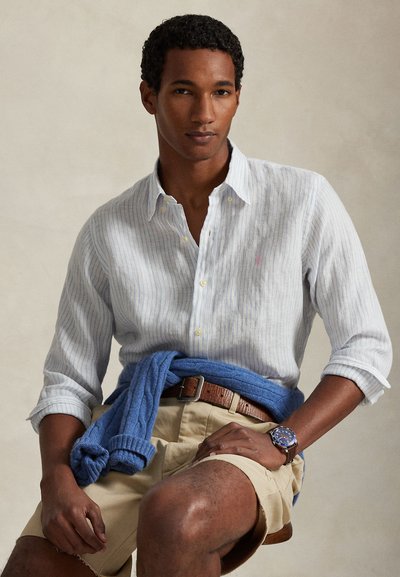 Πουκάμισο με γαλάζιες και λευκές ρίγες, με γυρισμένα μανίκια, συνδυασμένο με μπεζ σορτς. Μπλε πουλόβερ δεμένο γύρω από τη μέση, καφέ ζώνη και ασημένιο ρολόι.