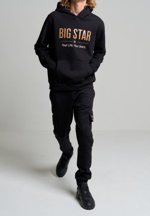 Osoba idąca w czarnej bluzie z kapturem z napisem "BIG STAR Your Life. Your Jeans.", dopasowanych czarnych spodniach typu cargo i czarnych tenisówkach.