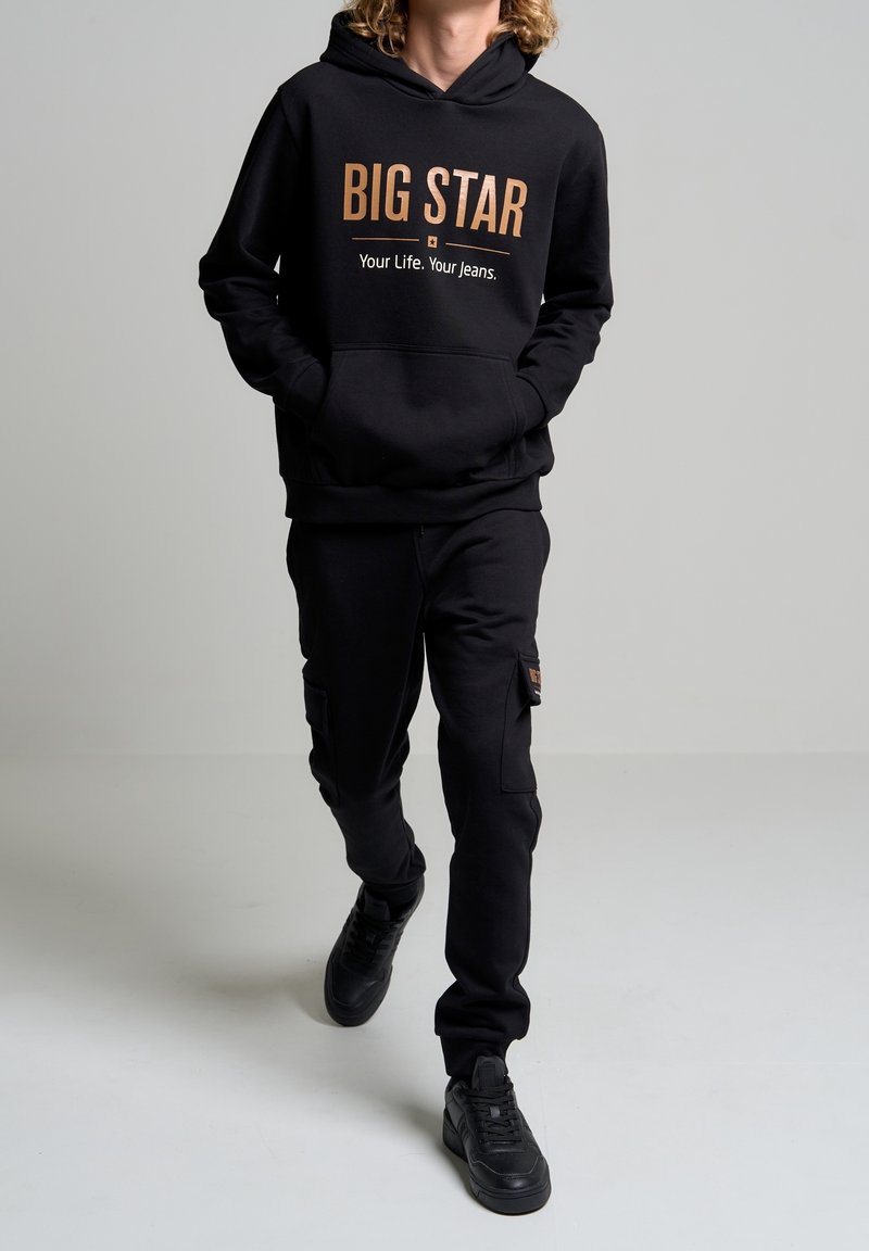 Personne marchant portant un sweat à capuche noir avec le texte « BIG STAR Your Life. Your Jeans. » et un pantalon cargo noir assorti ainsi que des baskets noires.