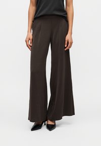 Γυναίκα που φοράει σκούρα καφέ παντελόνια με φαρδιά μπατζάκια και μαύρες μυτερές γόβες, εμφανιζόμενη από τη μέση μέχρι τα πόδια σε απλό φόντο.