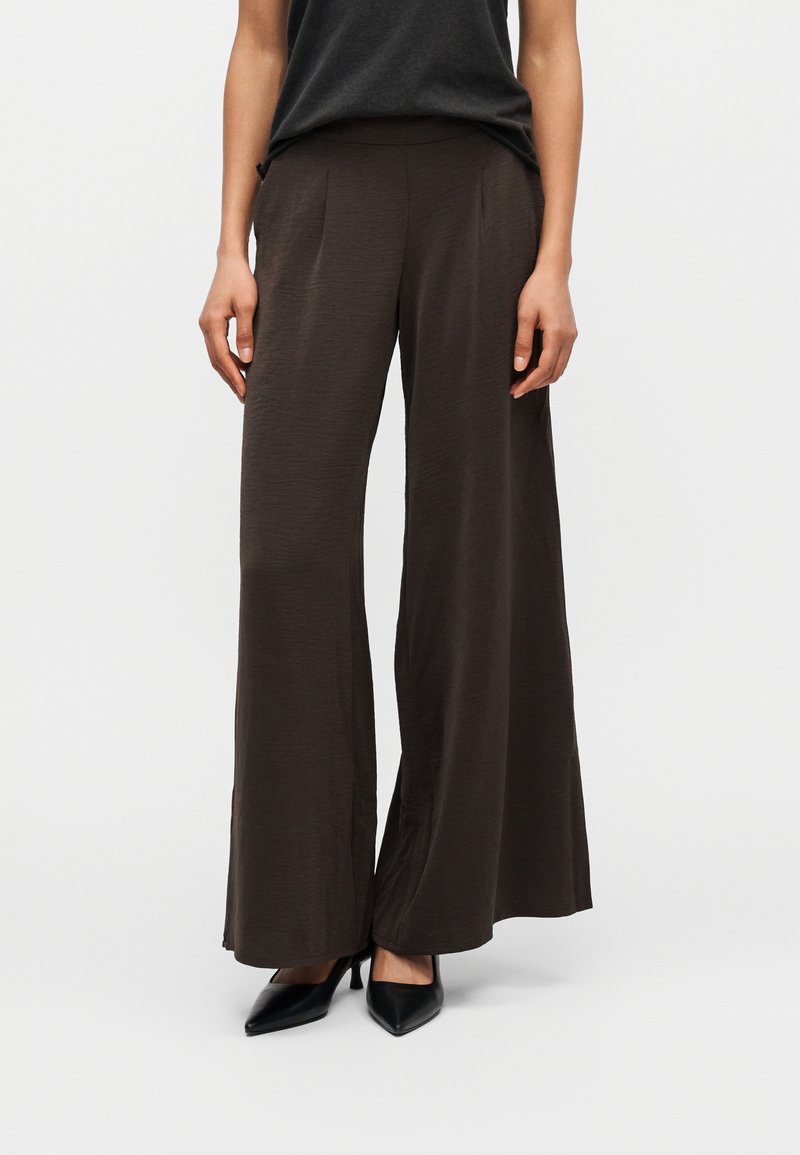 Γυναίκα που φοράει σκούρα καφέ παντελόνια με φαρδιά μπατζάκια και μαύρες μυτερές γόβες, εμφανιζόμενη από τη μέση μέχρι τα πόδια σε απλό φόντο.