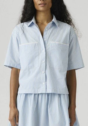 Kobieta ubrana w koszulę na guziki z krótkim rękawem w jasnoniebiesko-białą kratkę oraz dopasowaną spódnicę, na jednolitym tle.