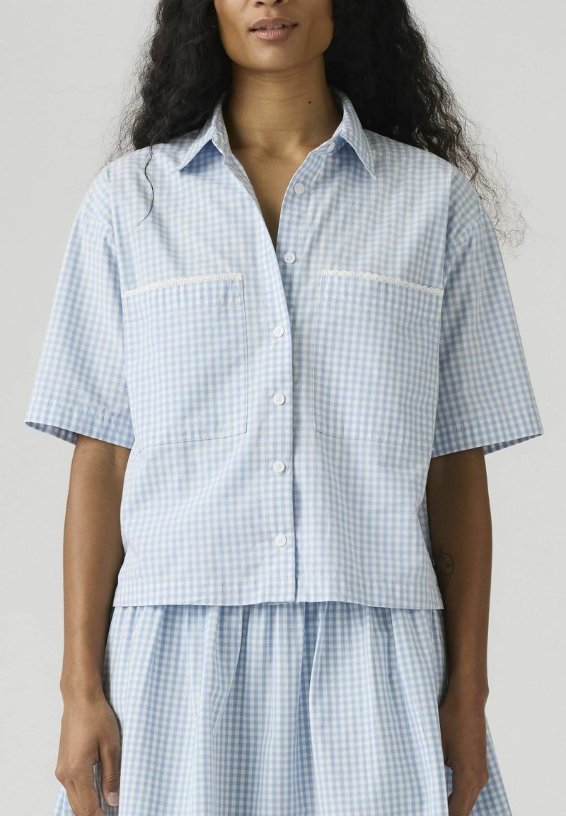 Femme portant une chemise à manches courtes boutonnée en vichy bleu clair et blanc avec deux poches avant, assortie à une jupe, sur fond uni.