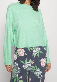Kobieta w miętowym swetrze z długim rękawem i granatowych spodniach z dużymi różowymi, białymi i zielonymi wzorami kwiatowymi.