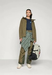 Giacca impermeabile verde oliva, t-shirt blu navy, camicia a quadri legata in vita, pantaloni khaki, porta uno zaino a fantasia, sneakers di colore chiaro.