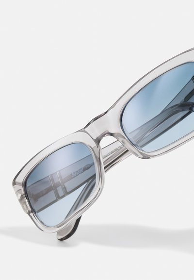 Óculos de sol transparentes com lentes azuladas, uma armação retangular espessa e uma textura suave. Apresenta um logótipo impresso no interior.