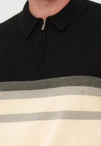 Μαύρο πλεκτό μπλουζάκι με γιακά και ημιδιακόρυφη κλείσιμο με φερμουάρ, με οριζόντιες ρίγες σε σκούρο γκρι, ανοιχτό γκρι και κρεμ στο ύψος του στήθους.