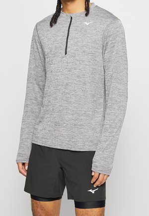 Persona che indossa una maglia sportiva grigia a manica lunga con cerniera frontale e pantaloncini neri a strati con loghi sul petto e sulla gamba.