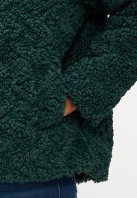 Zöld, bolyhos fleece dzseki textúrált felülettel, nagyméretű ujjakkal és elülső zsebbel. Puha, meleg anyag, ami ideális a hétköznapi viselethez.