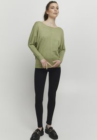 Világoszöld, laza szabású pulóver széles nyakkivágással és hosszú ujjakkal, fekete, testhezálló leggings-szel és fekete loaferekkel párosítva.