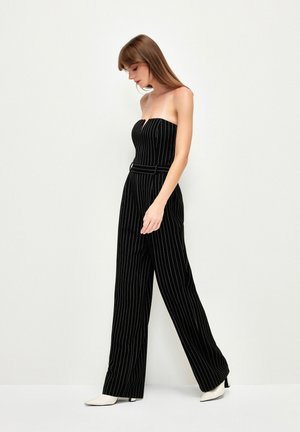 Mono negro con rayas, con parte superior sin mangas, piernas anchas y cintura ceñida con cinturón. Tela suave con un ligero brillo, acompañado de zapatos blancos de punta.