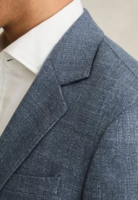 Hall muut tekstuuriga bleiser, millel on nurgapealsed läbib lapid, rindade tasku ja all valge ülikonna särk. Kangas on kudumismuster.