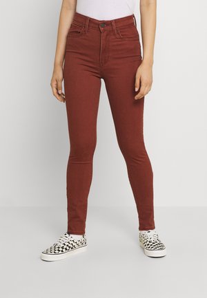 Femme portant un jean skinny taille haute couleur rouille avec poches avant, assorti à des baskets slip-on à carreaux noir et blanc.